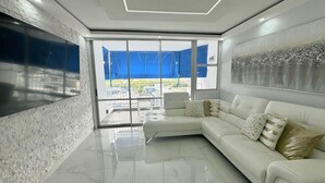 A modern living room with a white leather sofa, a glass coffee table, and a large flat-screen TV mounted on the wall. - Beachfront  2 Bdrm ~ Walk to Eat ~ W/D ~ King Bed (Carolina)