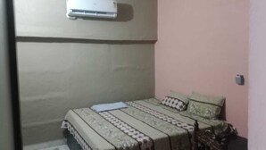 A small, single-bed room with a patterned bedspread, a wall-mounted air conditioner, and a plain wall. - HOLKAR SEVA SADAN (Mathura)