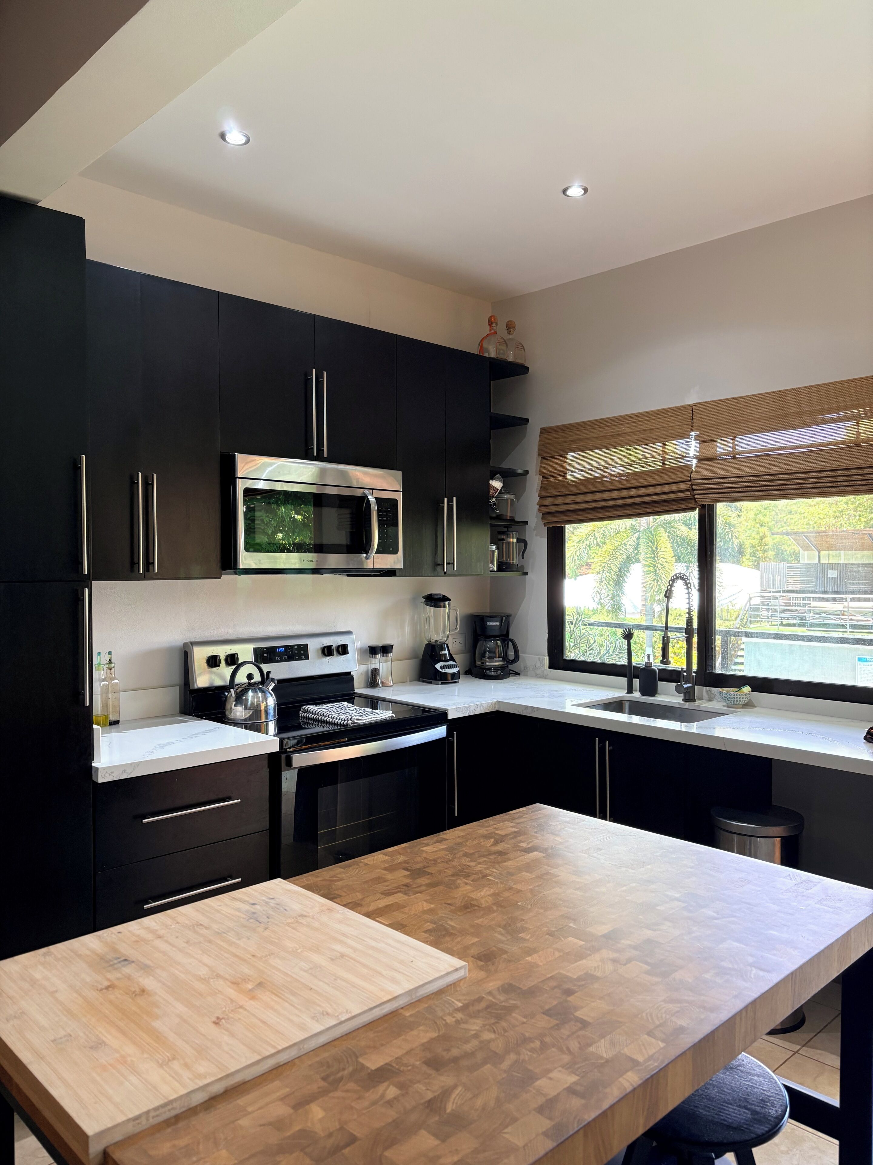A modern kitchen with dark cabinets, stainless steel appliances, a wooden countertop, and a window with a view of greenery.
