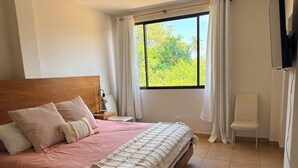 A bedroom with a bed, a nightstand, a chair, a window with curtains, and a TV mounted on the wall. - Coco Beach Costa Rica, 2 Bedrm 2 Bath condo with beautiful outdoor space (Guanacaste Province)