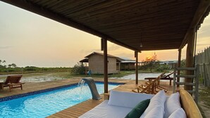 A wooden deck with a pool, a covered seating area with white cushions, and a view of the sunset over a distant building. - Aponem Spa Pousada (Prado)