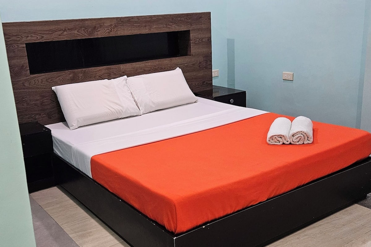 A bed with a white mattress and orange bedspread, a wooden headboard, and a pair of towels on the bed.