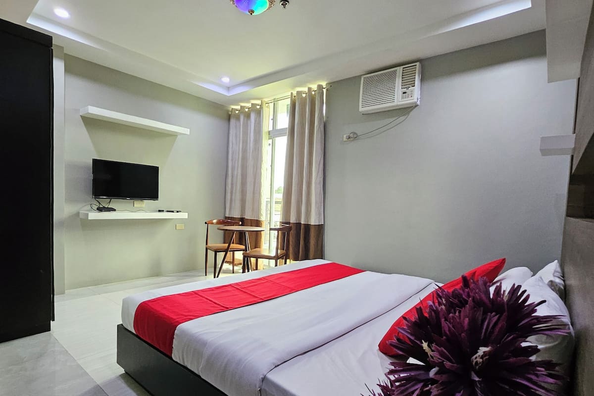 A modern hotel room with a bed, a TV, a small table with chairs, and a window with curtains.