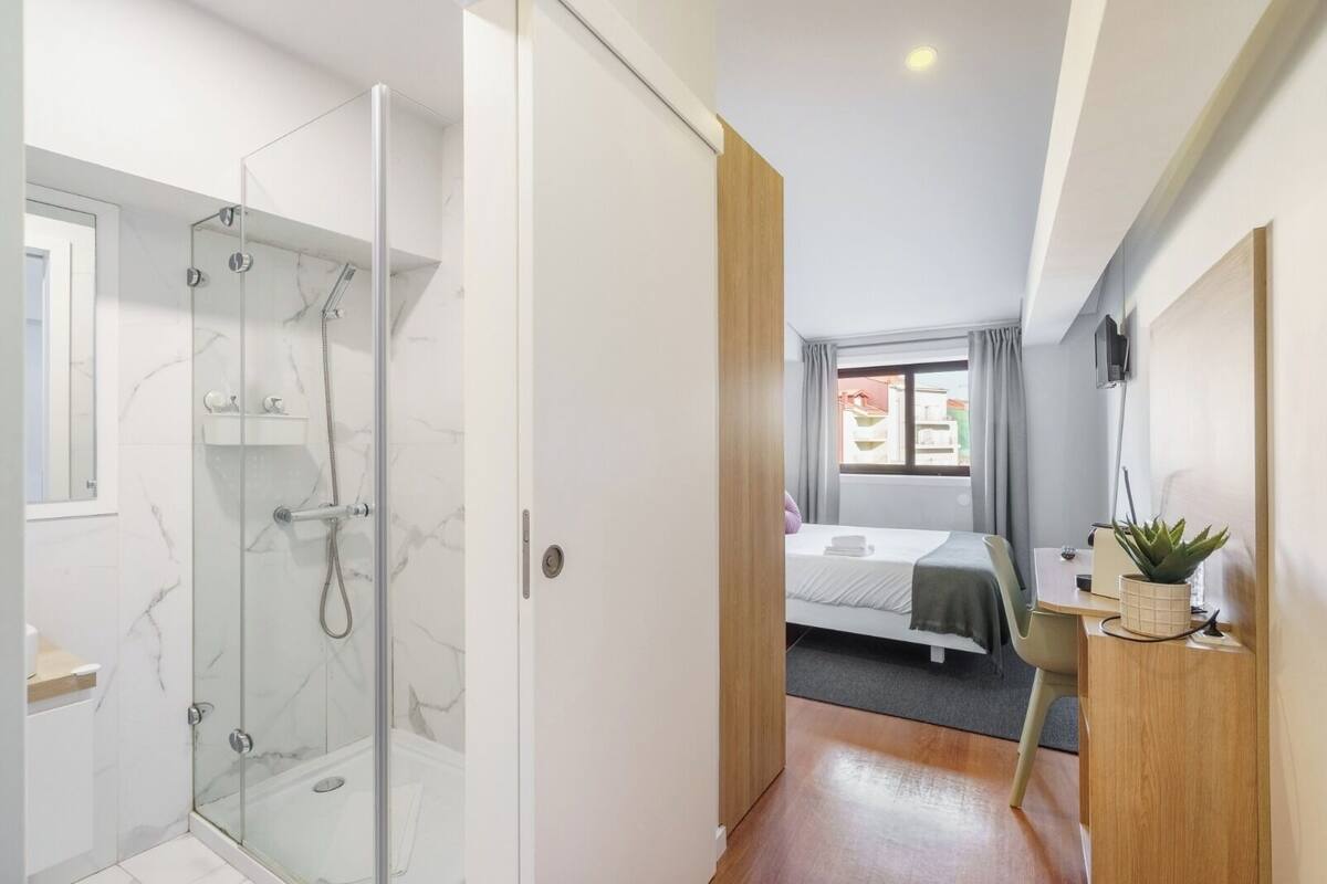 A modern hotel room with a glass-enclosed shower, a bed with a view of the city, and a small desk with a plant.