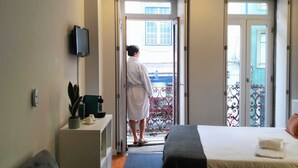 A hotel room with a bed, a desk with a plant, a TV, and a balcony with a railing. - Bellu Studios & Suites   (Braga)
