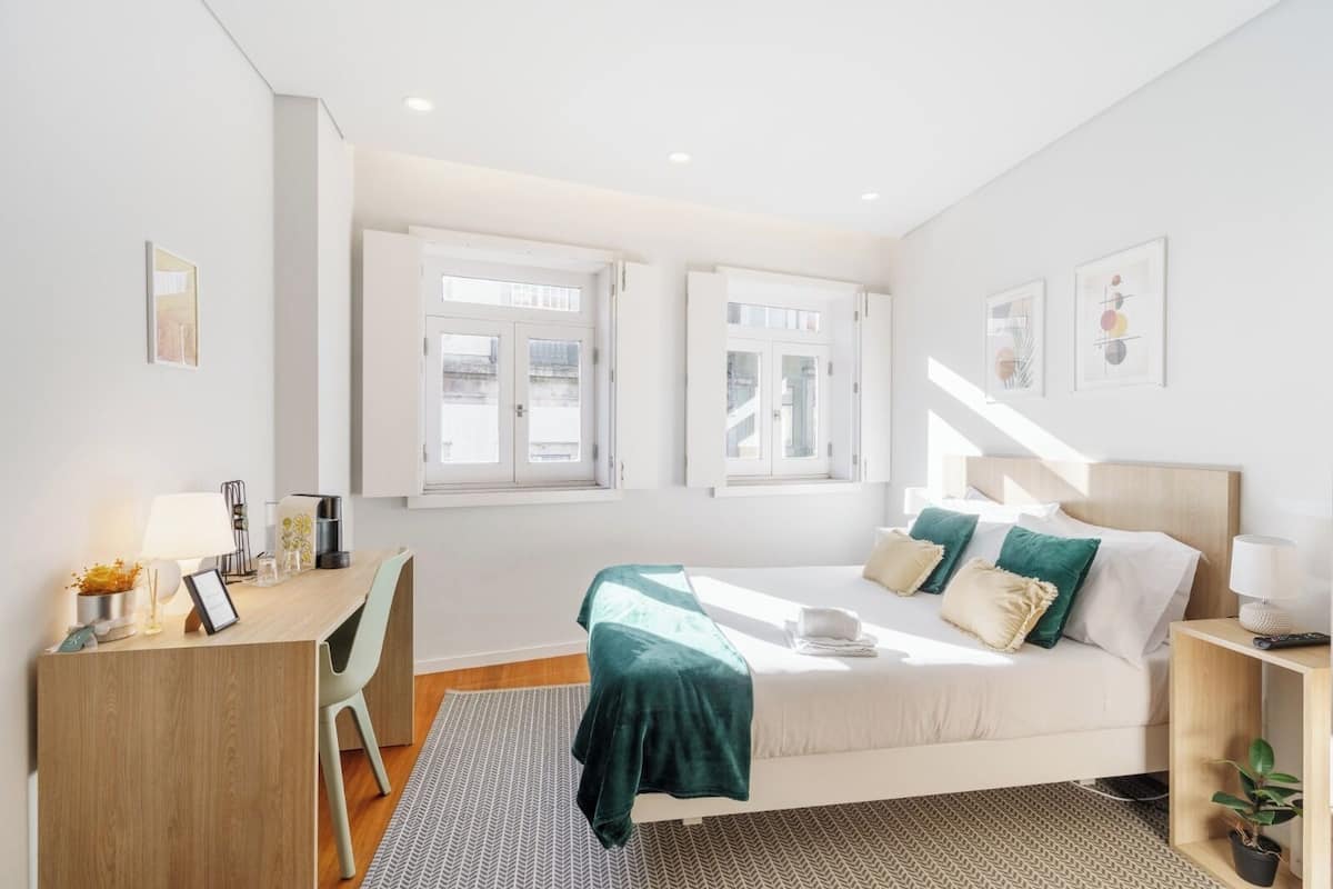 A modern bedroom with a bed, desk, and two windows.