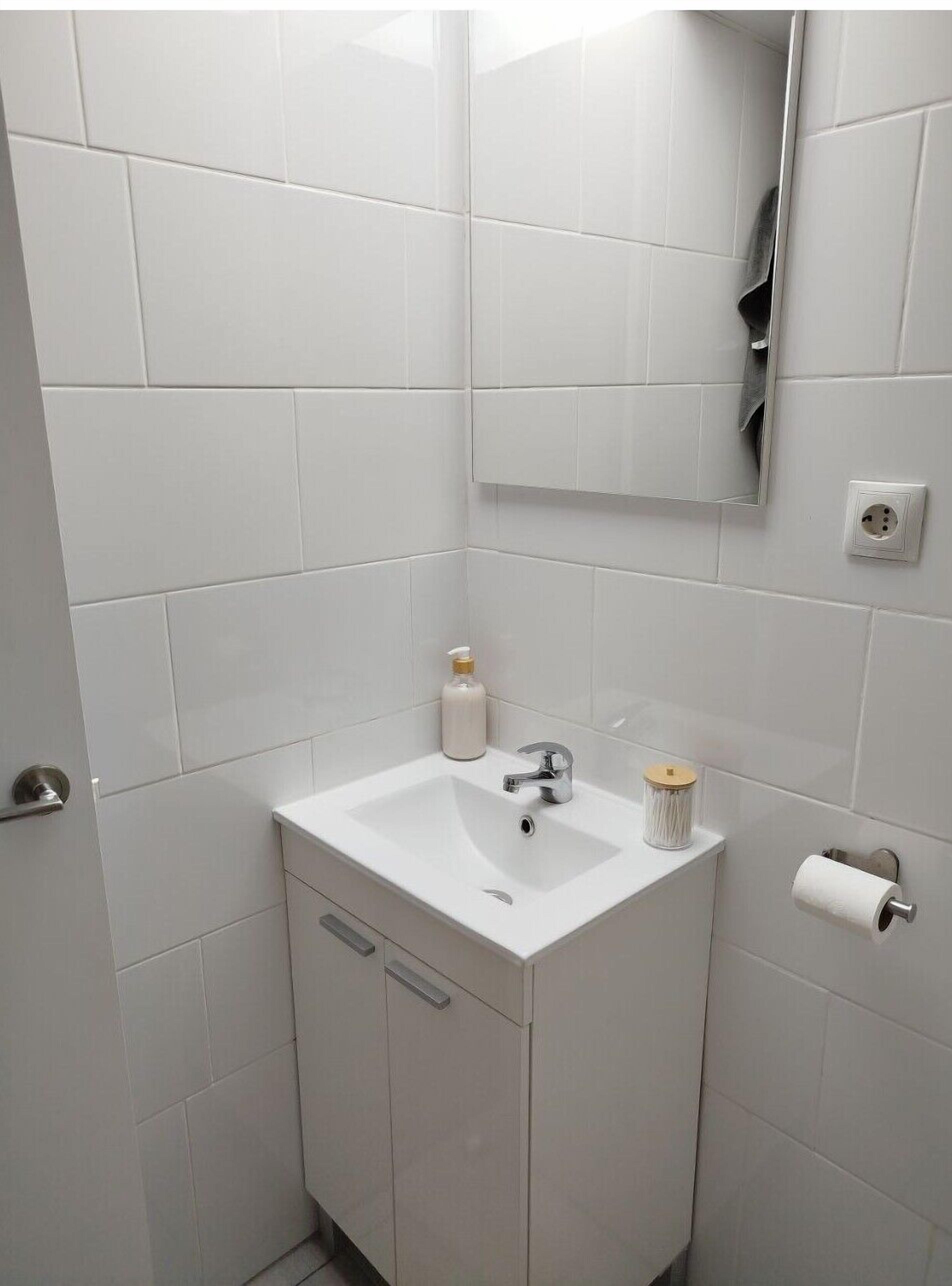 A bathroom with white tiles, a sink, a mirror, a soap dispenser, a toilet paper holder, and a towel dispenser.