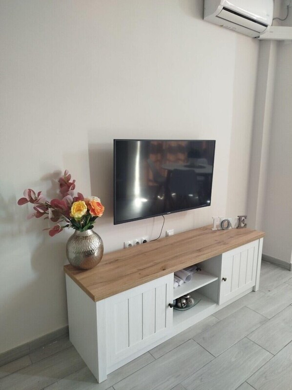 A modern living room with a flat-screen TV mounted on a white wall, a wooden console table with a vase of flowers, and a white cabinet below. - My Queen's Corner (Torremolinos)
