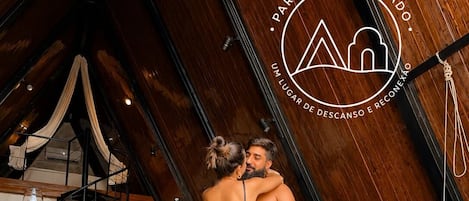 A couple embracing in a room with a high ceiling, a staircase, a dining area, and a leather sofa.