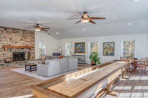 A spacious living room with a stone fireplace, a long wooden coffee table, a grey sofa, and a ceiling fan.