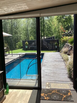 Zwembadgedeelte met een houten terras, een glazen deur en uitzicht op een tuin met bomen en een hek.