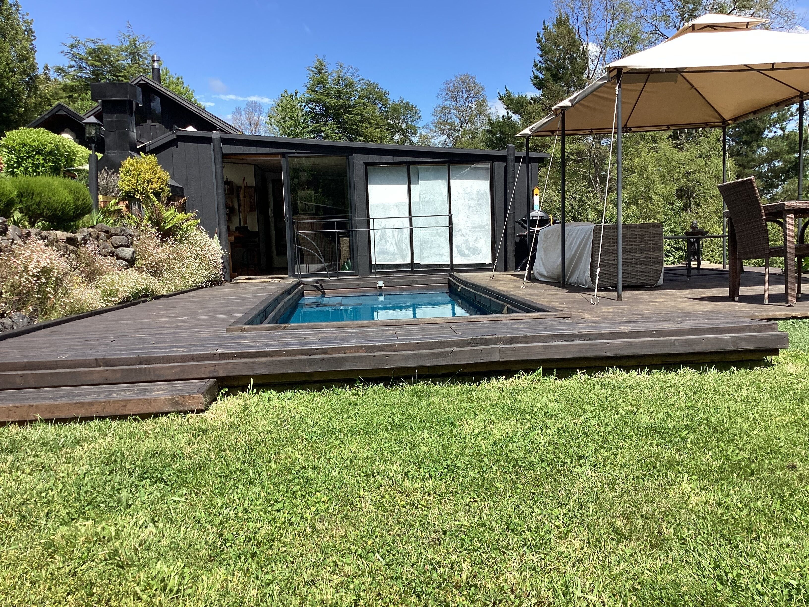 Un espace piscine extérieur moderne, doté d’une terrasse en bois, d’un coin salon abrité et d’une maison avec de grandes fenêtres.