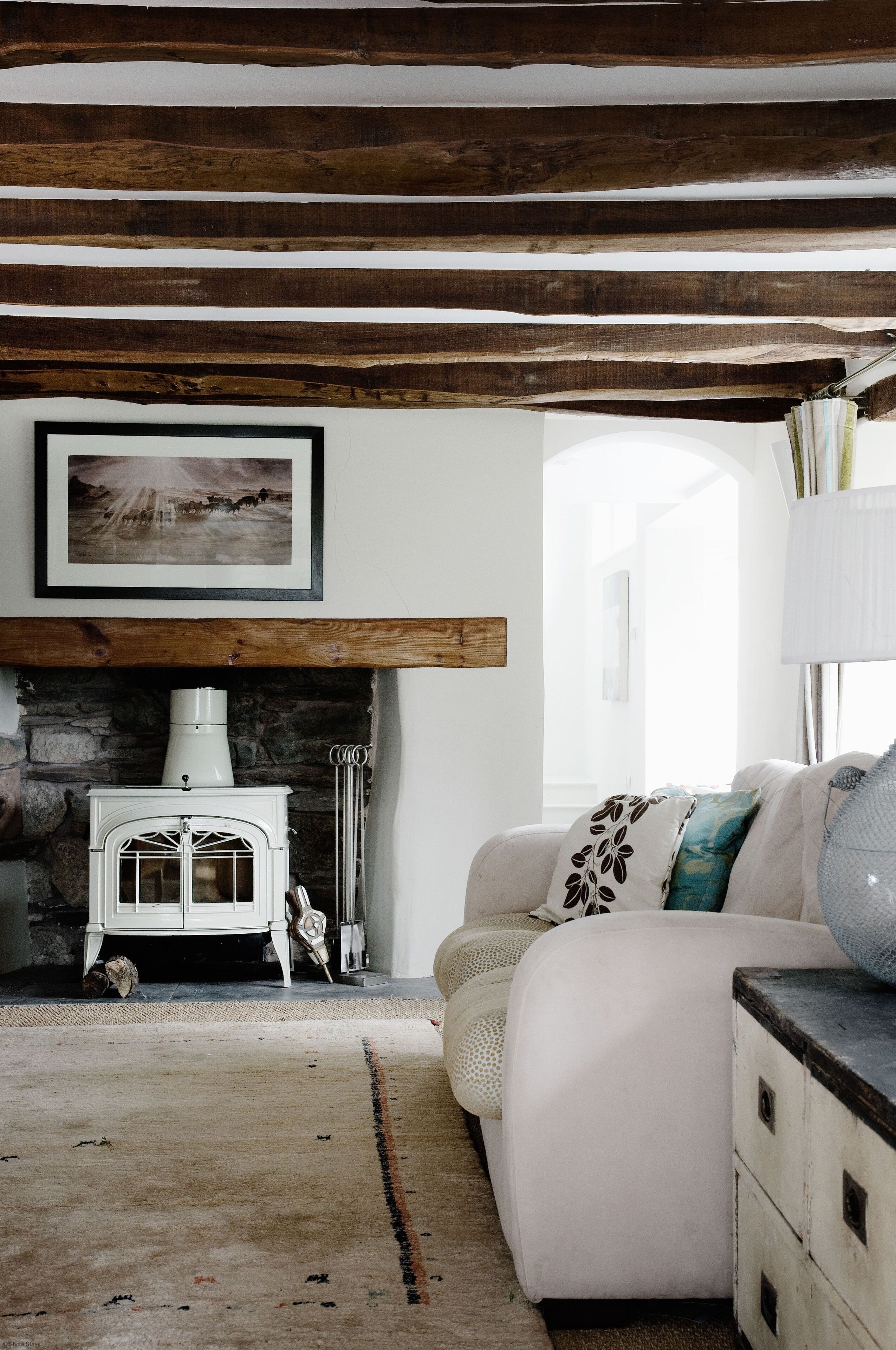 A cozy living room with exposed wooden beams, a white fireplace, a sofa, and a lamp.