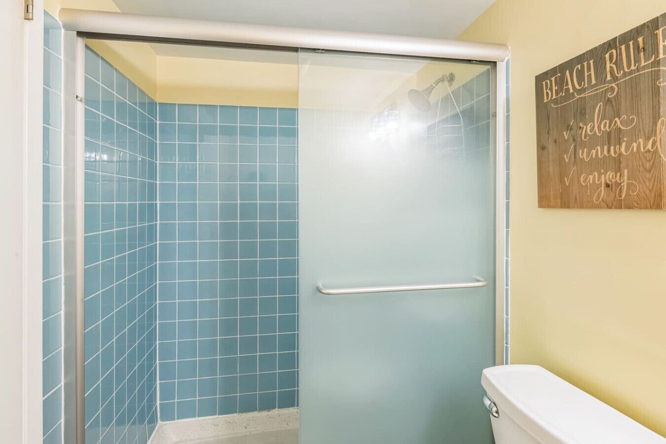 A bathroom with a glass shower door, blue tiles, and a wooden sign on the wall.