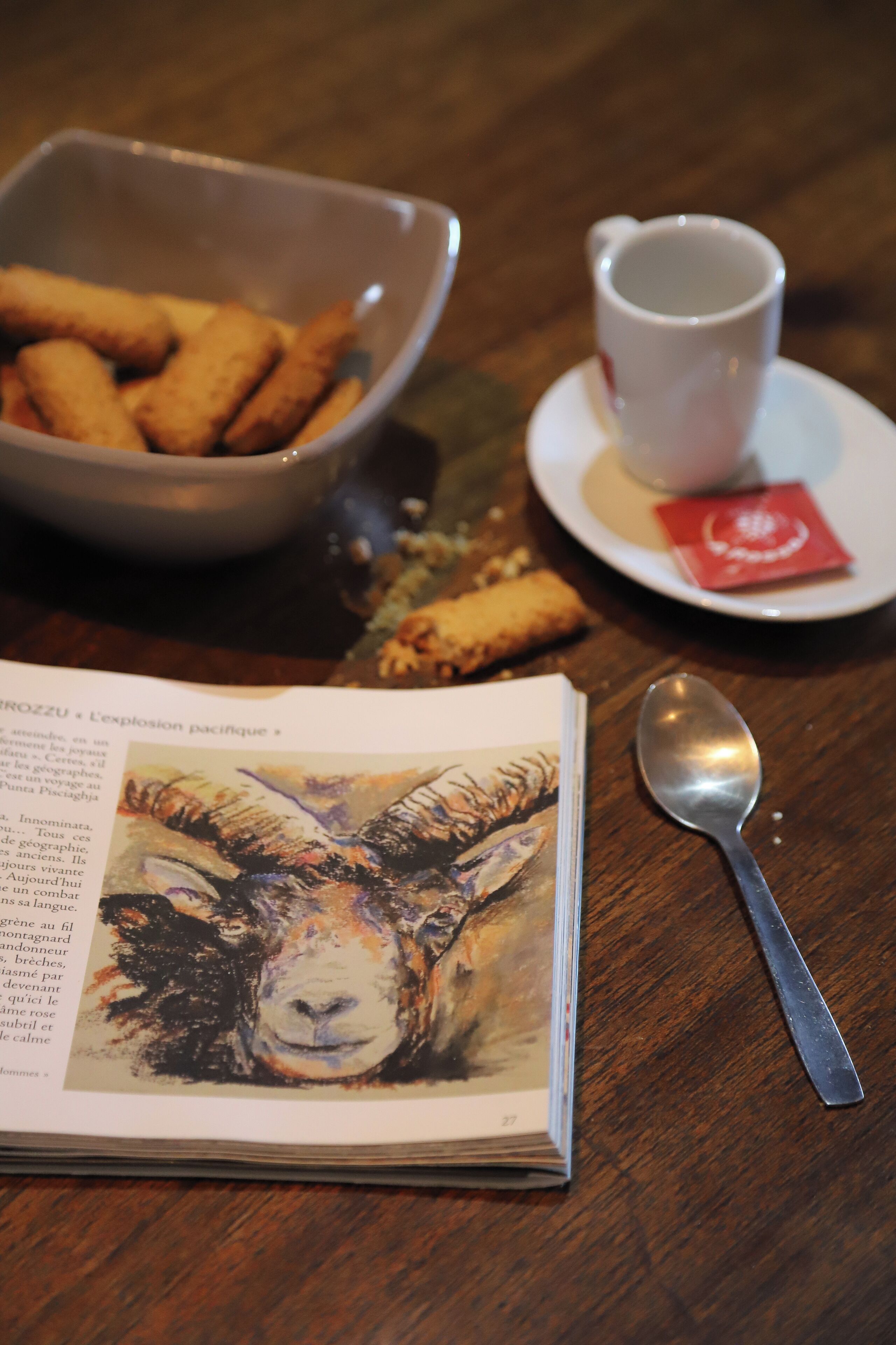 A table with a bowl of croquettes, a cup of tea, a spoon, and a book with a painting on the cover.