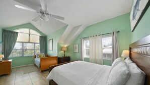 Ein Schlafzimmer mit einem Bett, einem Schreibtisch, einem Stuhl, einem Deckenventilator und einem Fenster mit Vorhängen.