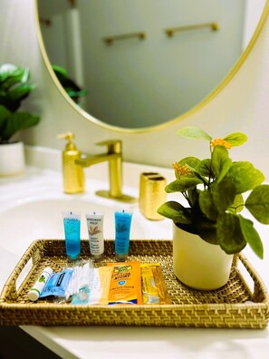 A bathroom with a white sink, a gold faucet, a round mirror, and a wicker tray holding toiletries including Bergman Kelly shampoo, body lotion, and Banana Nut Ultra Sport lotion. - Charming home near the beach, downtown and main golf courses. (Myrtle Beach)