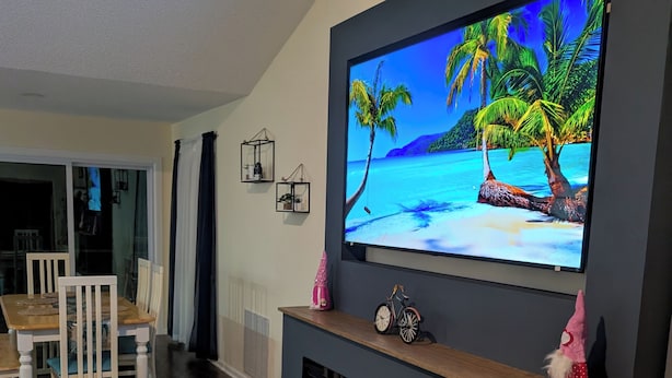 Una sala de estar moderna con un televisor de pantalla plana montado en una pared oscura, una mesa de comedor de madera con sillas y una chimenea con frente de vidrio.