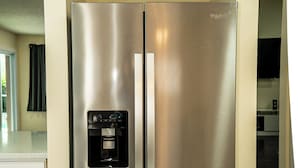 Un refrigerador Whirlpool de acero inoxidable con dispensador de agua, ubicado en una cocina con gabinetes blancos y piso de mármol.