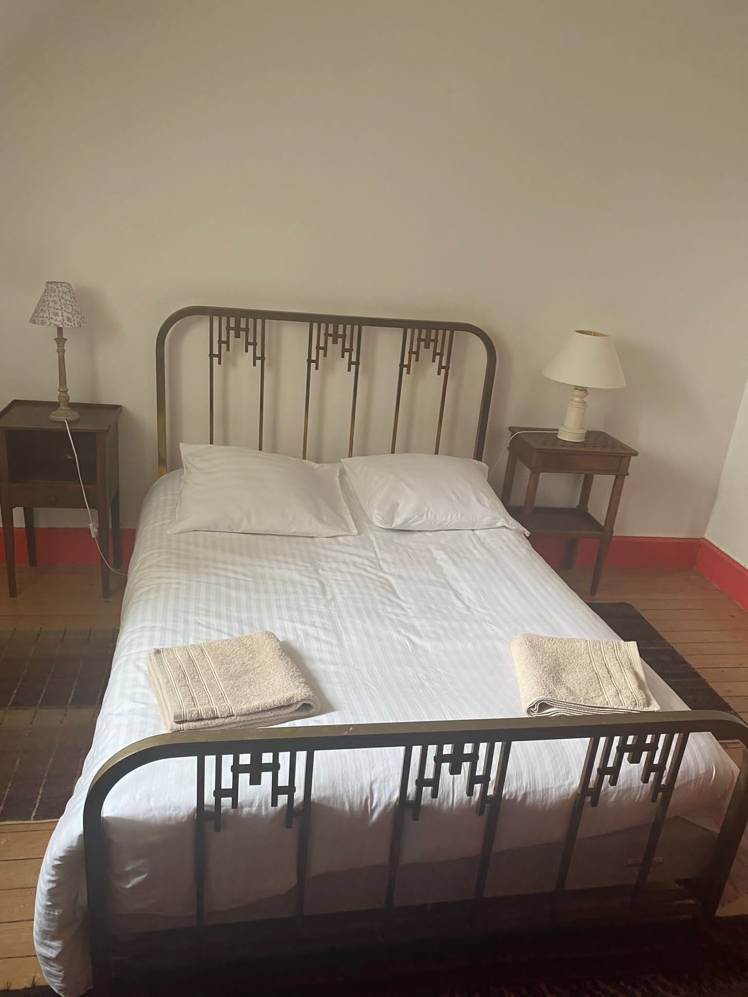 A bed with a metal frame, two pillows, and two folded towels. There are two bedside tables, each with a lamp. The room has a wooden floor and a plain wall.