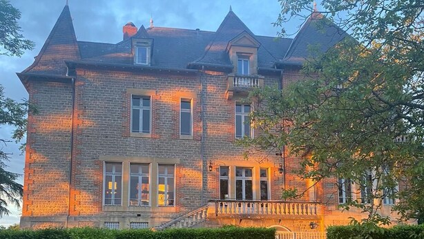 Un grand bâtiment en briques, à plusieurs étages, doté d’une façade symétrique, d’une entrée centrale avec des marches et d’une pelouse bien entretenue agrémentée d’une table et de chaises.