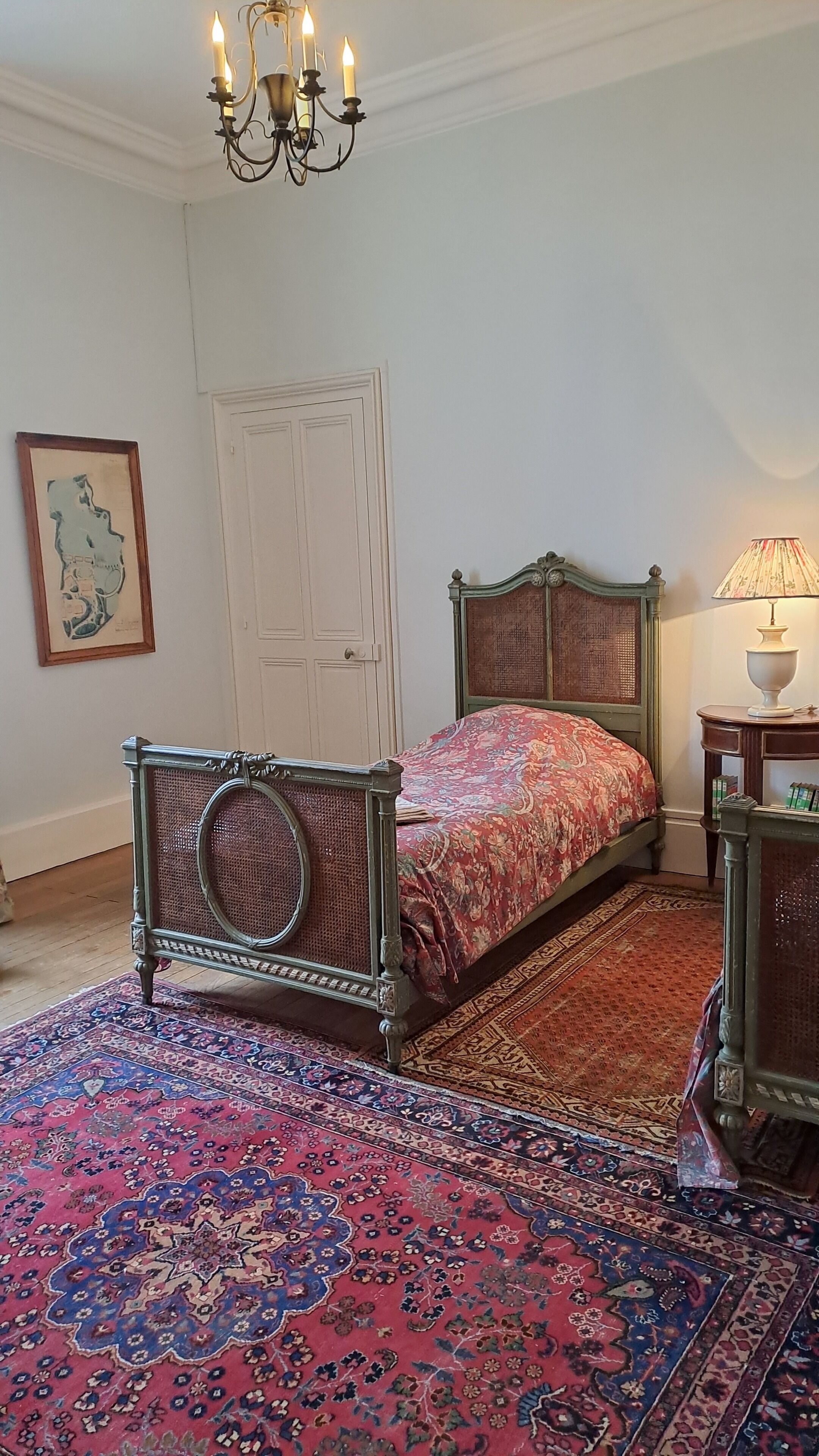 A bedroom with a bed, a chandelier, a lamp, a door, and a framed picture on the wall.