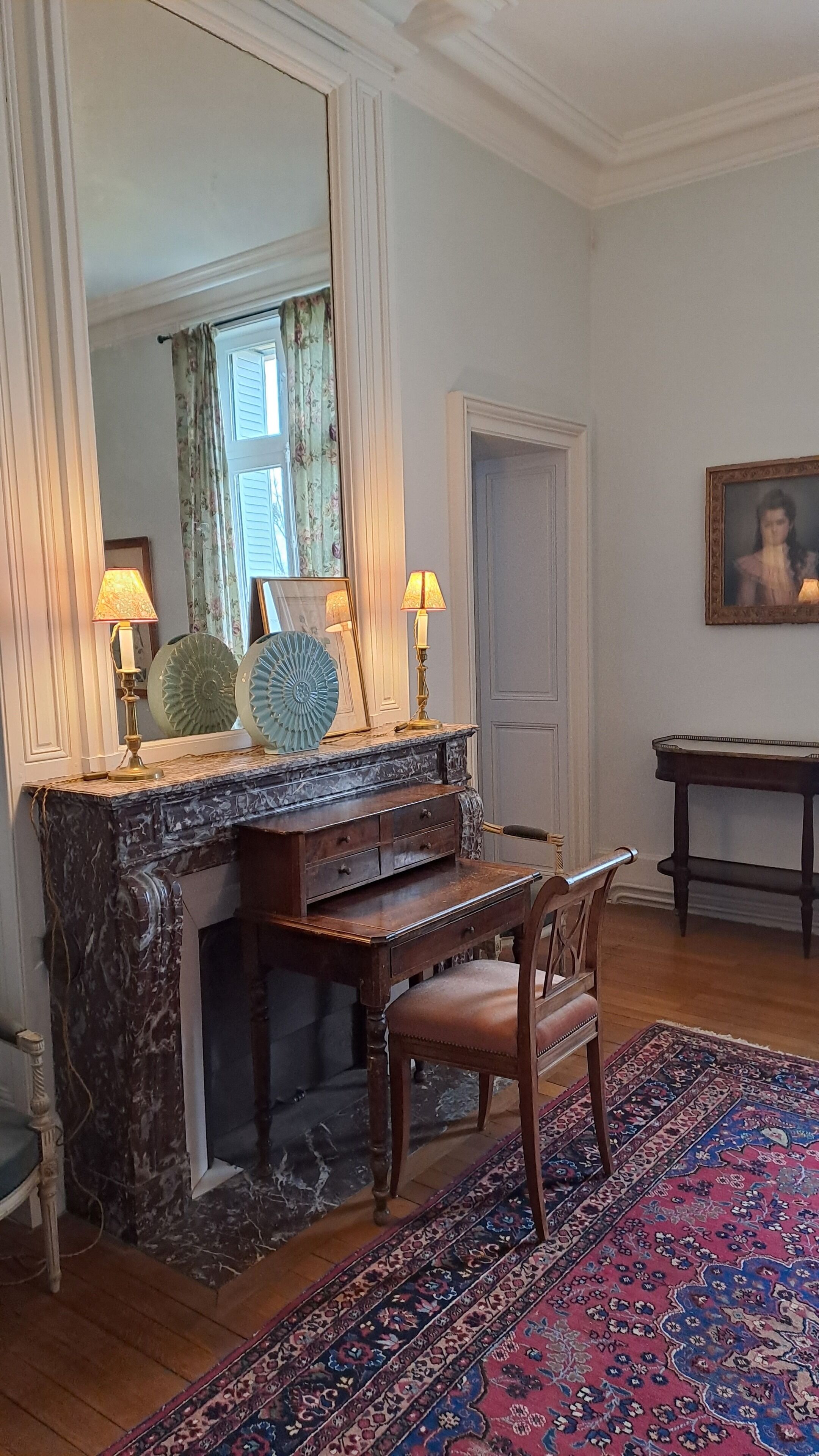 A room with a marble fireplace, a wooden chair, a large mirror, and a patterned rug.