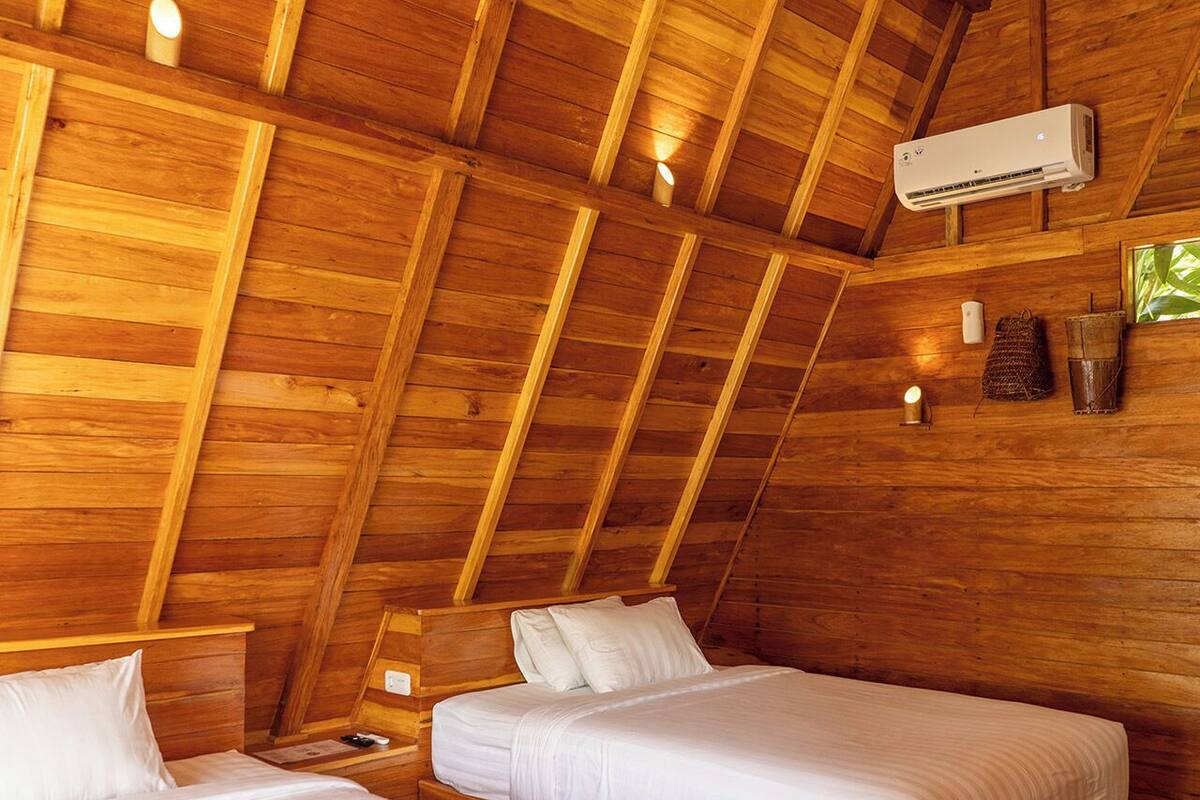 A cozy attic bedroom with wooden beams, two single beds, a wall-mounted air conditioner, and a small table with a lamp.