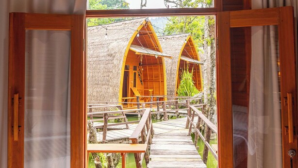A wooden bridge leading to thatched-roof cabins with a view of greenery and a wooden deck. - Torau Resort (Toinasa)