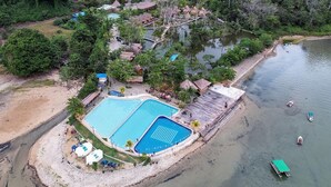 An aerial view of a resort with a large swimming pool, a beach area, and a forested backdrop. - Torau Resort (Toinasa)