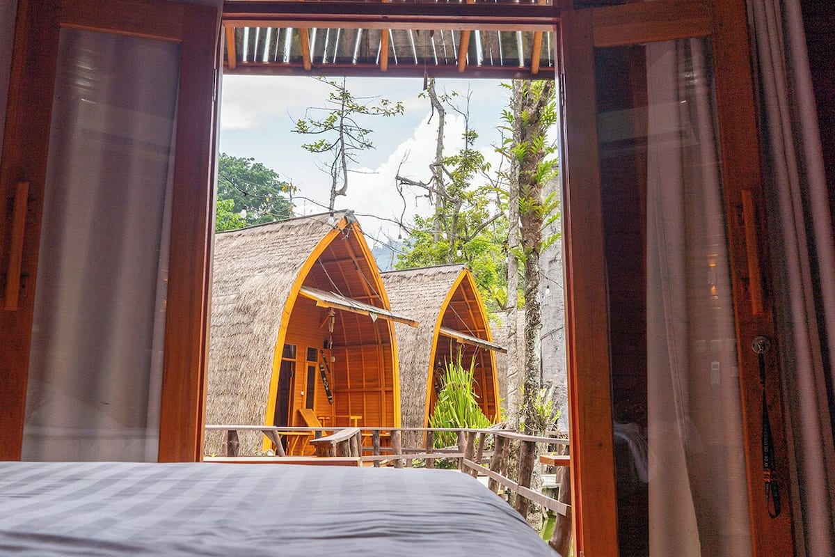 A room with a bed, wooden doors, and a view of two thatched-roof structures outside.