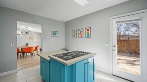 Una cucina moderna con isola blu, una zona pranzo con sedie arancioni e vista su un giardino retrostante tramite una porta in vetro.