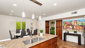 Une cuisine moderne dotée de plans de travail en granit, d’un évier en acier inoxydable, de meubles en bois, de luminaires suspendus et d’un coin repas avec une table et des chaises.