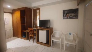 Een compacte kamer met een houten garderobe, een kleine witte koelkast, een televisie op een houten standaard en twee witte plastic stoelen.