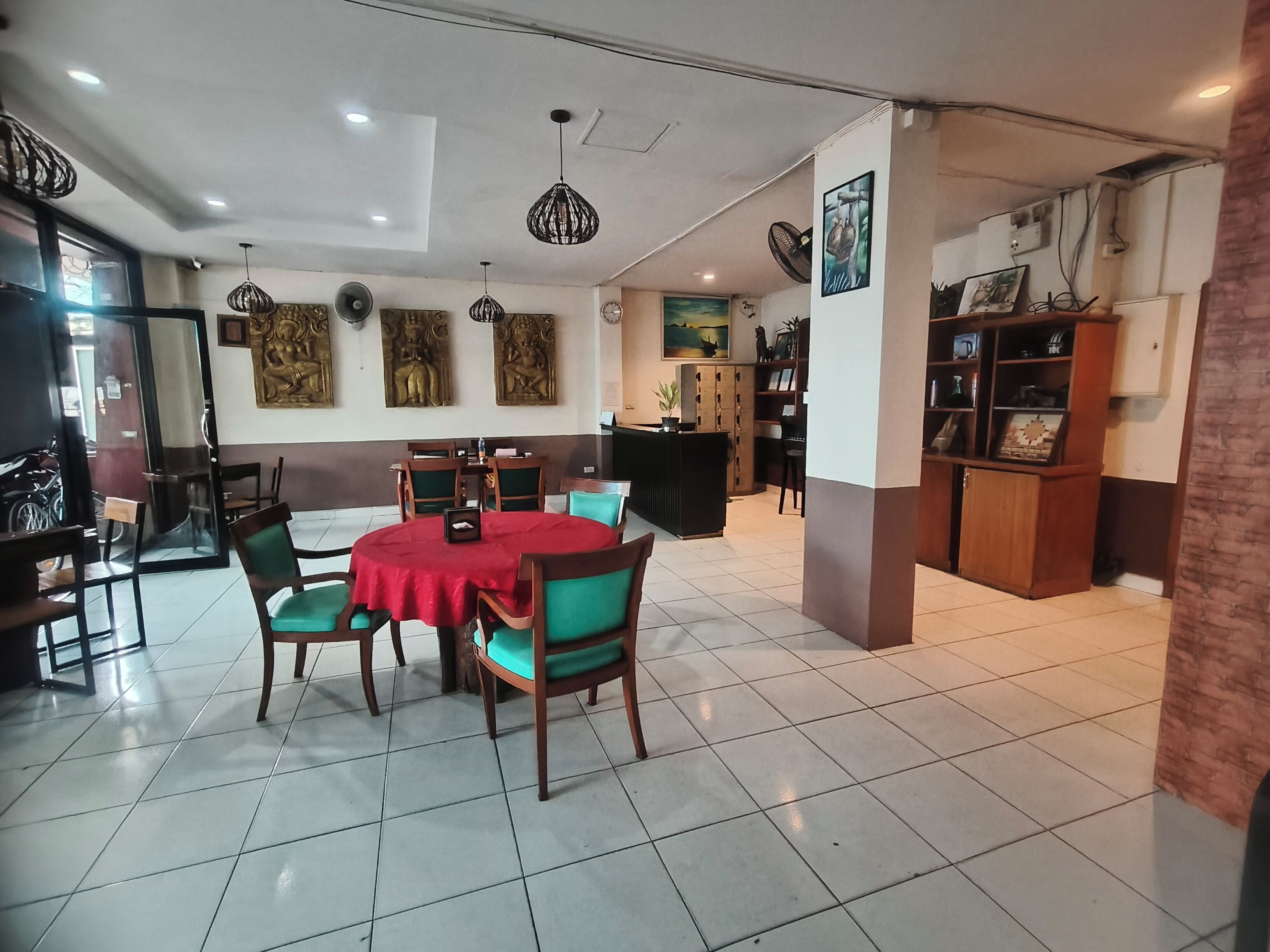A dining area with a round table covered in a red tablecloth, surrounded by wooden chairs with green cushions. There are ceiling lights and wall-mounted decorations.