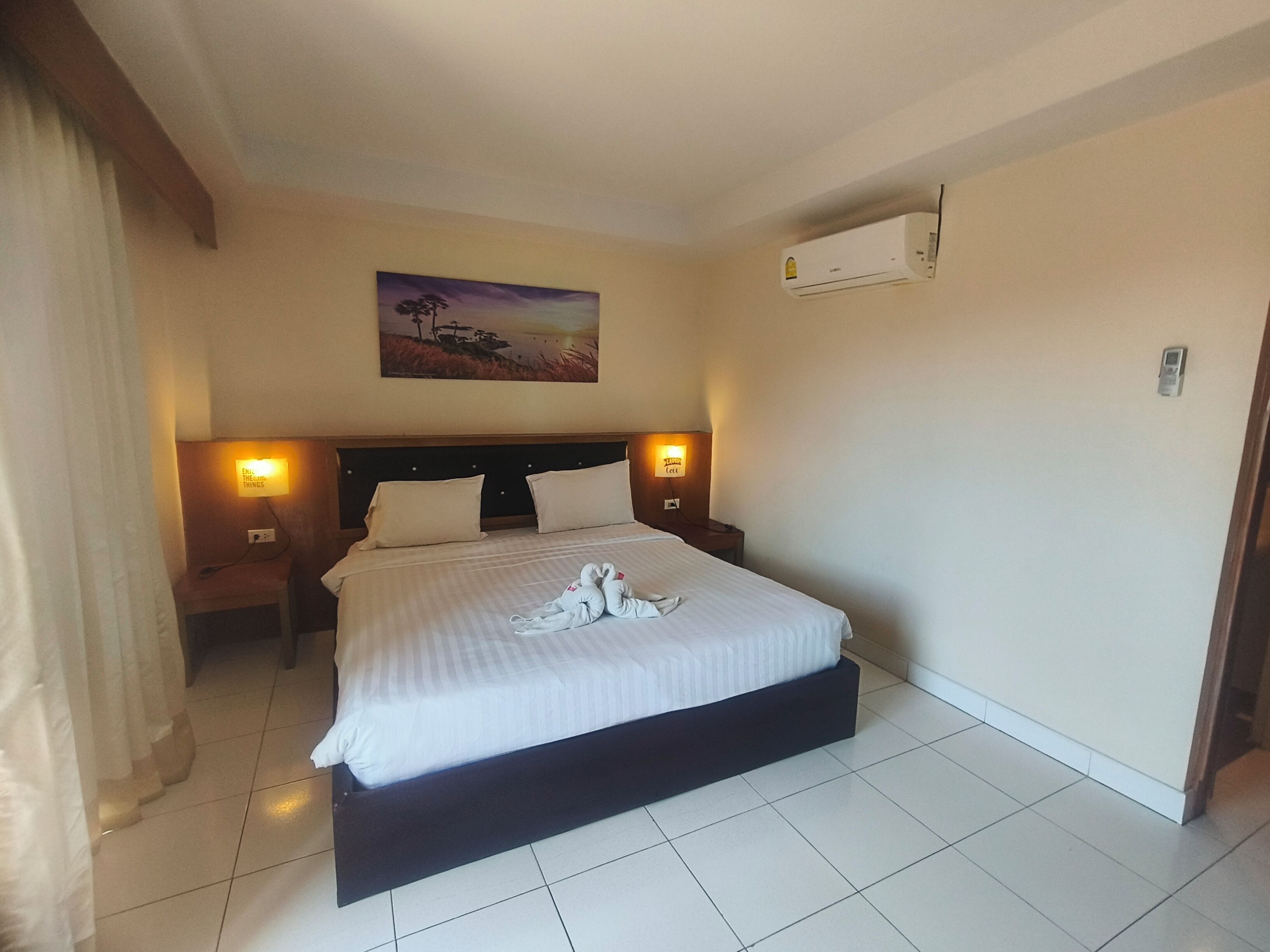 A hotel room with a large bed, two bedside lamps, a painting on the wall, and an air conditioning unit.