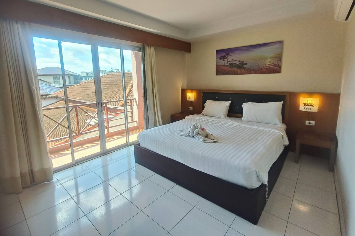 A hotel room with a large bed, two bedside tables with lamps, a painting on the wall, and a view of buildings through a sliding door.