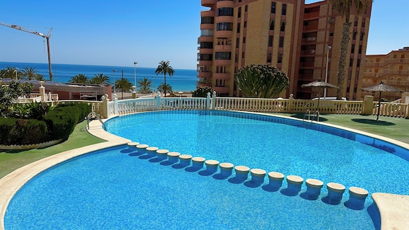 A swimming pool with a white deck and a white fence, surrounded by greenery and a view of the ocean.