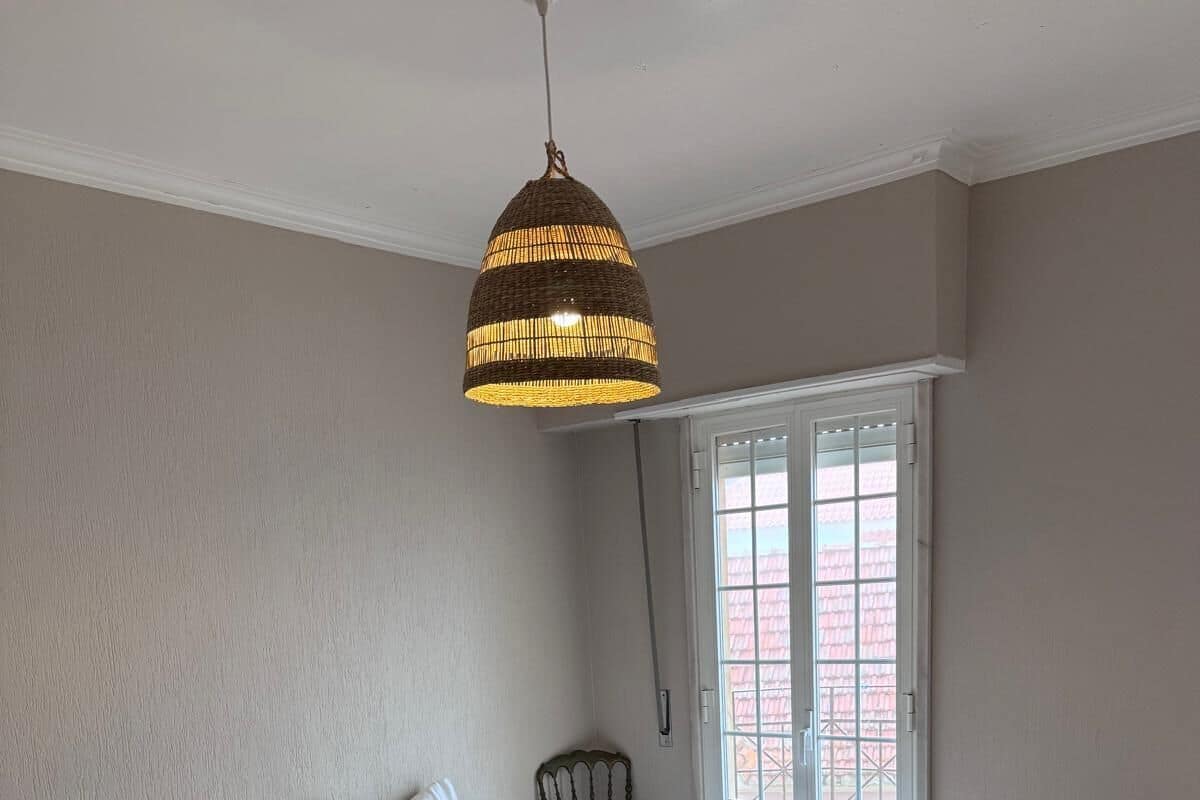 A bedroom with two beds, a hanging light fixture, a chair, and a window with a view of buildings.