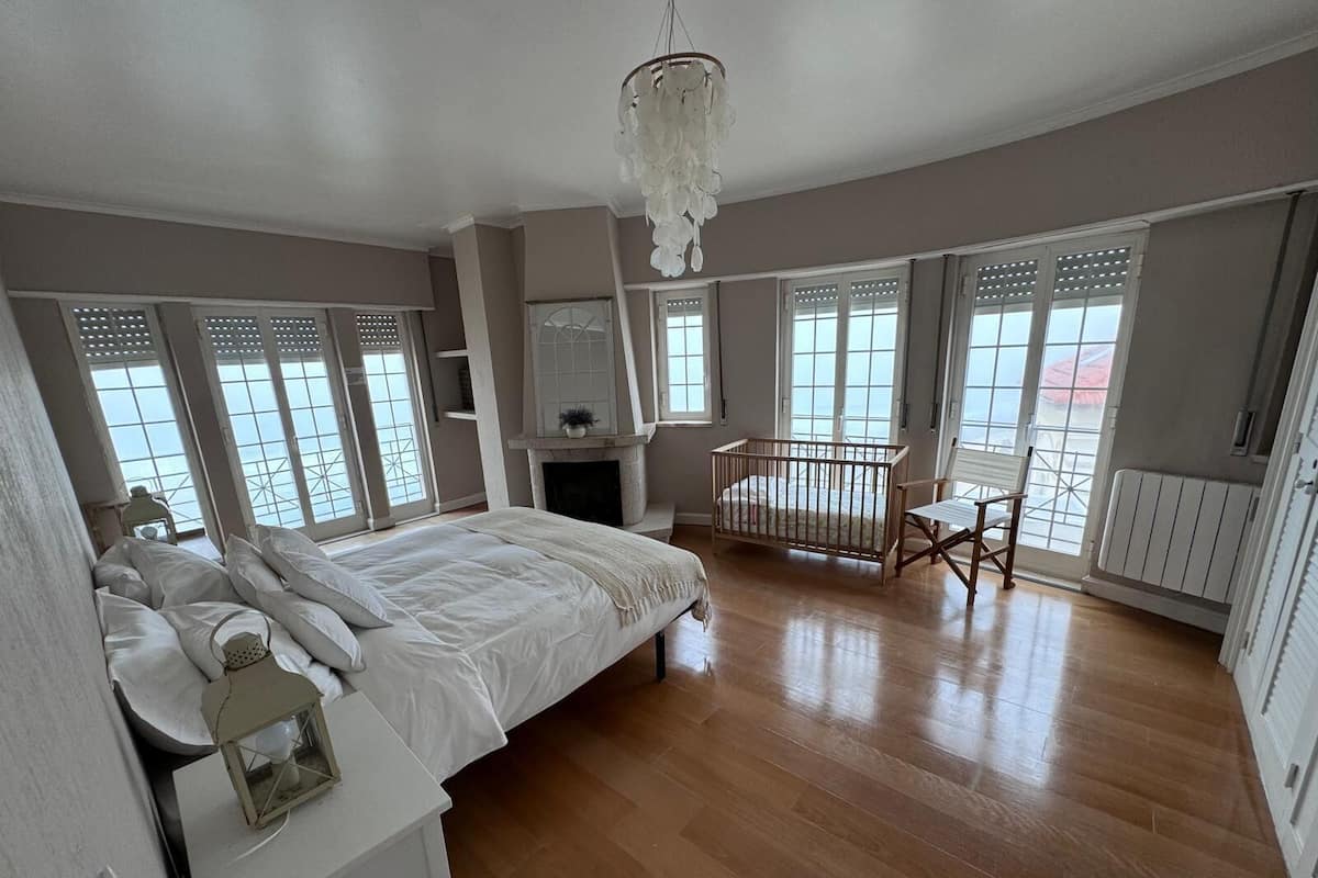 A spacious bedroom with a large bed, a crib, and a view of the sea through large windows.