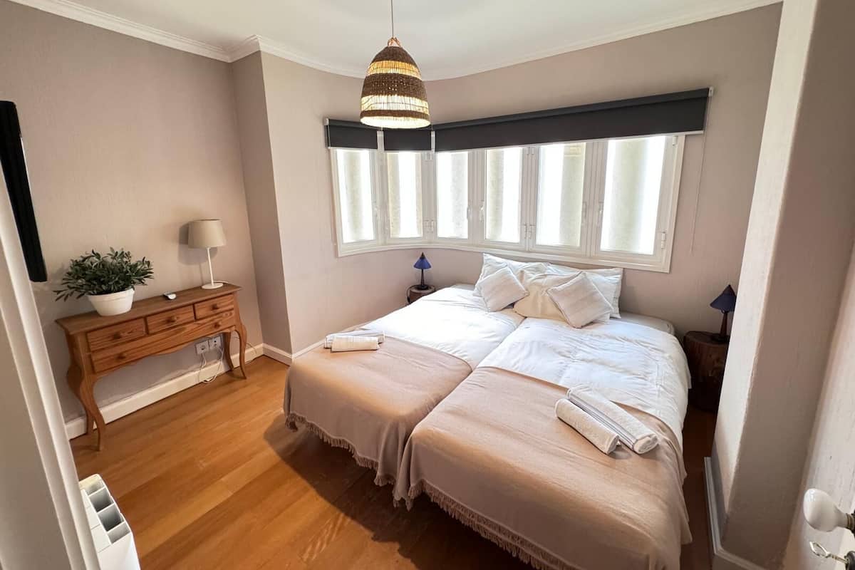 A bedroom with a double bed, a bedside table with a lamp, a dresser, a potted plant, and a window with blinds.