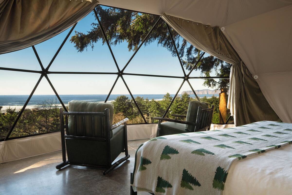 A tent with a bed and two chairs, a large window, and a view of the ocean.