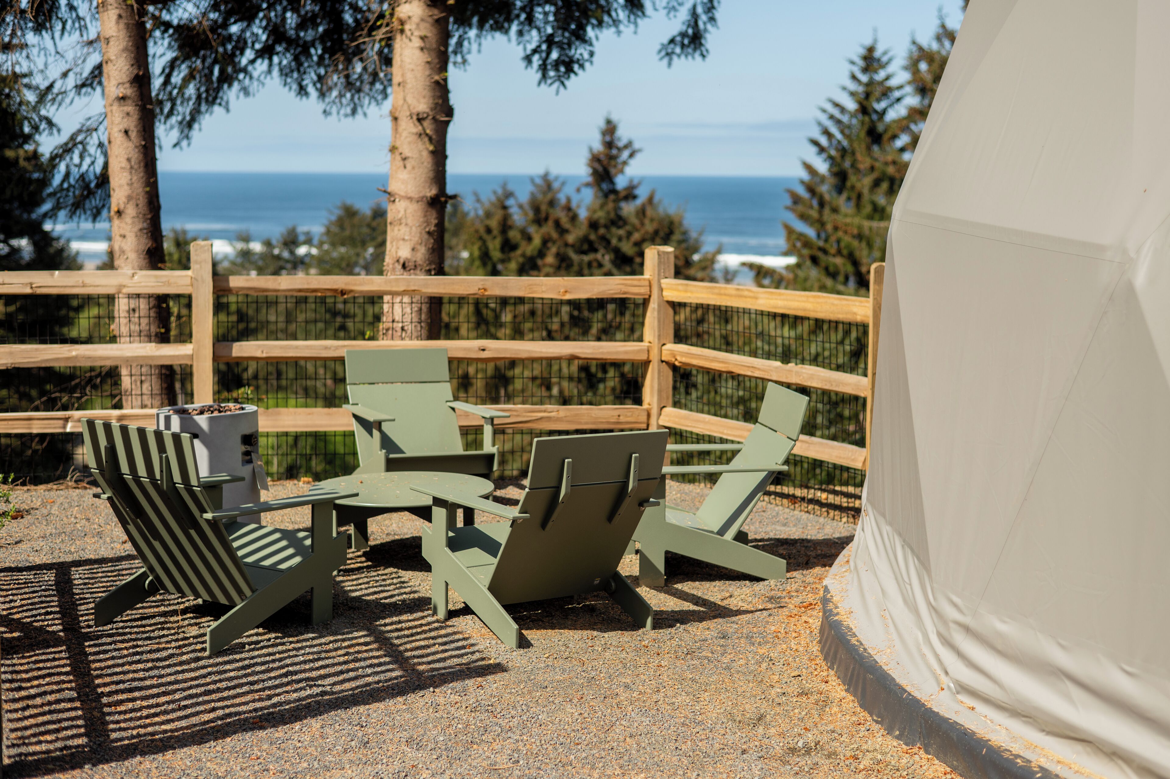 A seating area with Adirondack chairs and a table, a white tent, and a view of the ocean.