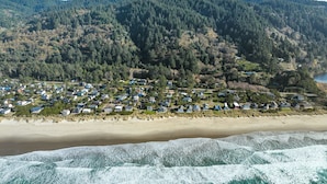 A coastal residential area with houses along the beach, a forested hill in the background, and waves crashing onto the shore. - Two Capes Lookout (Cloverdale)