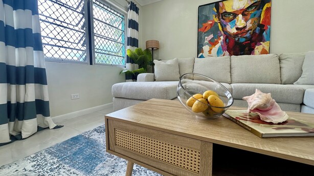 Una sala de estar con un sofá, una mesa de centro con un frutero, un cuadro grande y una ventana con cortinas.