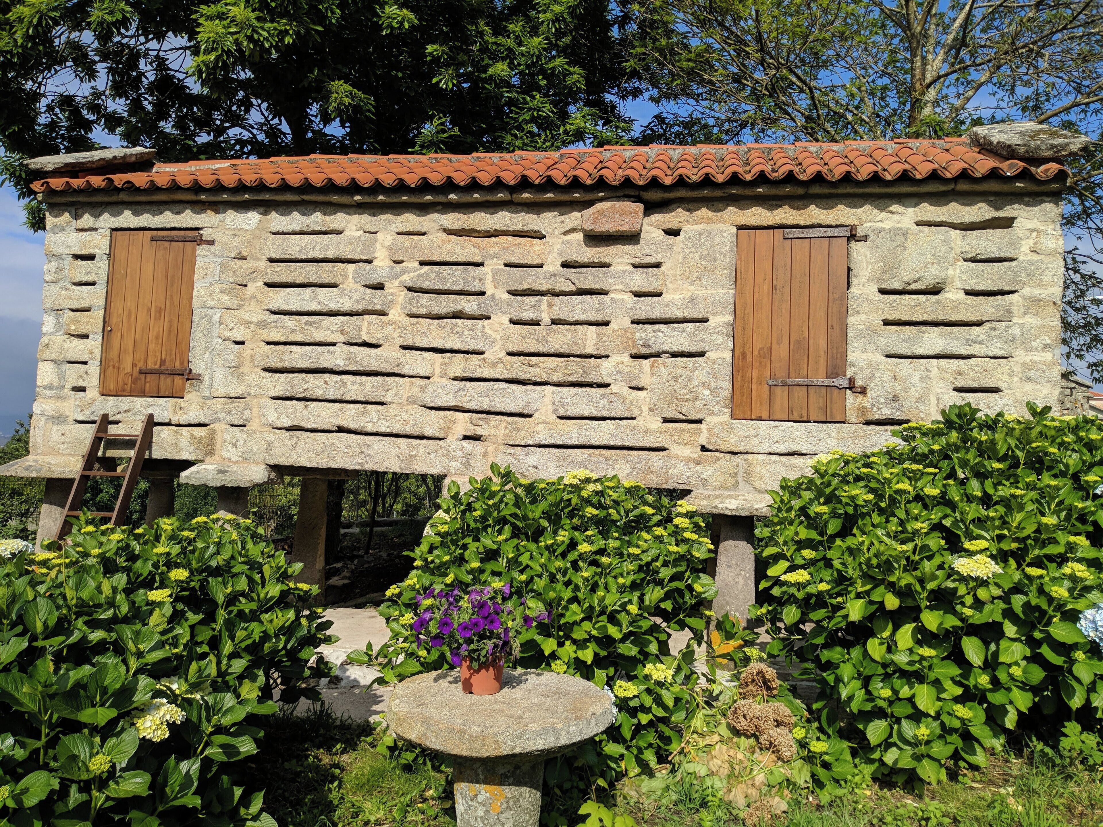 A stone building with a tiled roof, wooden shutters, and a stone bench with flowers.