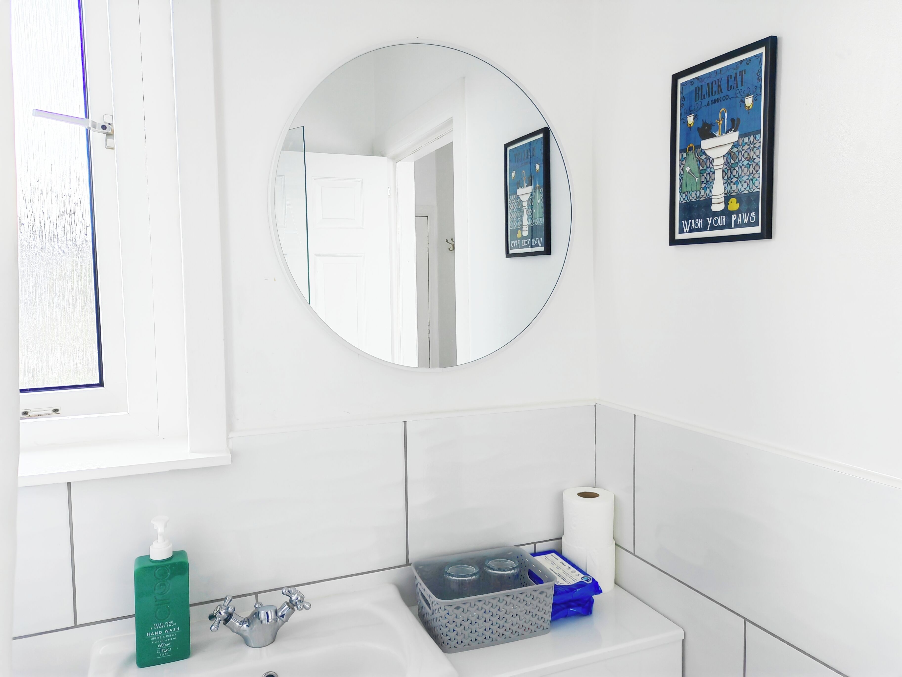 Une salle de bain avec un lavabo blanc, un miroir, un tableau encadré et une fenêtre avec des stores.