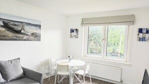 A room with a grey sofa, a white dining table with four chairs, a painting of a boat on the wall, and a window with a view of greenery. - Lovely flat near University of Aberdeen (Aberdeen)