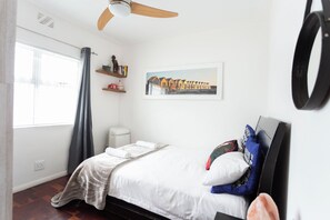 A bedroom with a bed, a ceiling fan, a window with curtains, a wall-mounted shelf with books and decorative items, and a framed picture on the wall.
