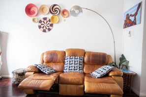 A leather sofa with patterned cushions, a floor lamp, and decorative woven items on the wall.